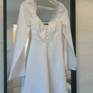 Vit långärmad klänning Gina Tricot - Supersöt vit klänning från Gina Tricot med lång ärm, markerad midja och knappar framtill. Klänningen har en lätt strukturerad yta, fyrkantig ringning och smock i ryggen för extra komfort. Perfekt för dig som gillar romantisk och stilren look. Endast använd en gång. Finns inte att köpa längre.