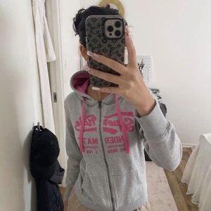 Grå hoodie med rosa tryck från MED-UPSTAR - Grå hoodie med dragkedja från MED-UPSTAR. Stor rosa text på bröstet, rosa snören och insida av huvan. Praktiska fickor framtill och ribbade muddar. Materialet är 60% bomull och 40% polyester. Perfekt för dig som gillar en sportig och chill stil.