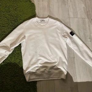Vit Stone Island sweatshirt - Vit sweatshirt från Stone Island med klassisk rund hals och ribbade muddar. Tröjan har den ikoniska svarta patchen med logga på vänster ärm. Tillverkad i mjuk bomull och har en clean, stilren look som passar perfekt till jeans eller cargobyxor.