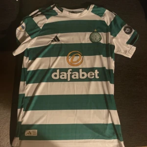 Celtic FC randig matchtröja Adidas - Celtic FC officiell matchtröja från Adidas med gröna och vita breda ränder, klubbmärke på bröstet och sponsorlogga. Korta ärmar, lätt och ventilerande material perfekt för fotboll. Märkt med William Hill Championship och Celtic FC Foundation på ärmarna.