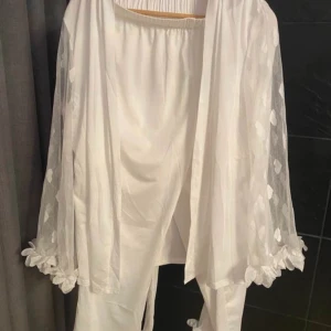 Vit omlottkostym med hjärtmönster - Säljer en vit tvådelad kostym med omlottkavaj och byxa. Kavajen har transparenta ärmar med hjärtmönster och fina volangdetaljer vid ärmslut. Byxan har elastisk midja och matchande volang vid benslut. Materialet känns mjukt och flowy, perfekt för dig som gillar romantiska detaljer.