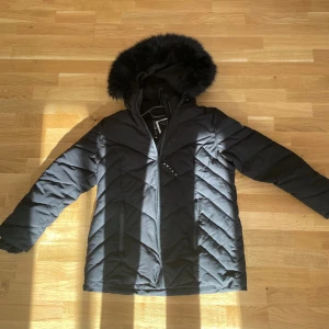 Svart pufferjacka från Amisu med fuskpäls - Svart pufferjacka från Amisu med snyggt quiltat mönster och huva med fluffig fuskpäls. Jackan har dragkedja framtill och två praktiska fickor med dragkedja. Perfekt för kalla vinterdagar och riktigt skön att bära.