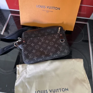 Svart axelväska monogram - Snygg svart axelväska från med klassiskt monogrammönster i grått. Väskan har justerbar axelrem, silverfärgade metalldetaljer och dragkedja. Tillverkad i skinn med präglad logotyp på både väska och tillhörande liten ficka framtill.