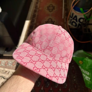 Rosa Gucci keps i jeans med GG-mönster - Säljer en rosa keps från Gucci i jeansmaterial med ikoniskt GG-mönster över hela kepsen. Kepsen har böjd skärm och en liten guldfärgad detalj på sidan. Perfekt för dig som vill sticka ut med en lyxig och trendig accessoar.