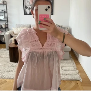 Ljusrosa volangblus med spetsdetaljer - Supersöt ljusrosa blus i tunn bomull med volanger längs axlar och ärmlös design. Blusen har v-ringning och fina spetsdetaljer framtill och baktill. Perfekt för dig som gillar romantisk och luftig stil.