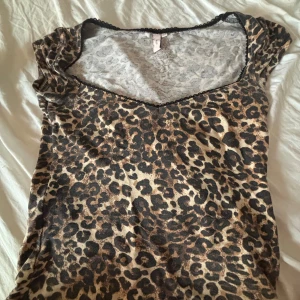 Leopardmönstrad topp med spetskant - Säljer en slim leopardmönstrad topp från H&M med v-ringning och svart spetskant. Toppen är kortärmad och har en figurnära passform. Perfekt för dig som vill sticka ut med ett djurmönster och gillar trendiga detaljer.