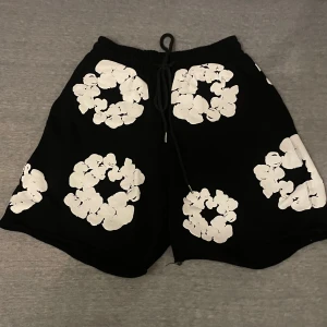 Svarta shorts från Denim Tears med blommor - Svarta shorts från Denim Tears med stora vita blommönster över hela plagget. De har elastisk midja med snörning och en loose fit. Perfekta för sommaren och riktigt snygga till streetwear-looken.