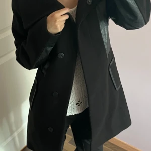 Svart kappa från H&M med skinnimitation - Säljer en svart kappa från H&M i storlek 34. Jackan har snygga detaljer i skinnimitation på axlar och fickor, samt knäppning framtill med stora knappar. Perfekt för dig som gillar en stilren och modern look.