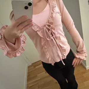 Rosa volangblus med knyt från Gina Tricot - Supersöt ljusrosa blus från Gina Tricot med volangdetaljer längs kanterna och vid ärmsluten i storlek XS. Blusen har långa ärmar och knyt framtill för en extra feminin touch.