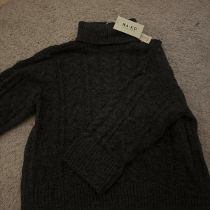 Mörkgrå polotröja från NA-KD - Mysig mörkgrå polotröja från NA-KD med kabelstickat mönster. Tröjan har hög polokrage, långa ärmar och breda ribbade muddar. Oanvänd med prislappen kvar.