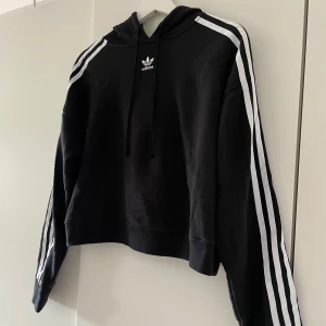 Svart Adidas hoodie  - Svart croppad hoodie från Adidas med klassiska vita ränder längs ärmarna och liten logga framtill. Tröjan har huva med snörning och långa ärmar. Perfekt för en sportig och chill stil.