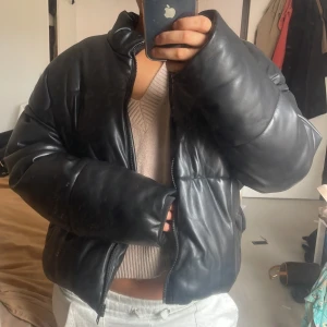 Svart pufferjacka i skinn-look - Säljer en svart pufferjacka med skinn-look och oversized passform. Jackan har quiltad design, dragkedja framtill och höga kragen. Perfekt för dig som gillar chunky och trendiga ytterplagg med street vibe. Jätteskön och varm. Nypris låg på 900kr