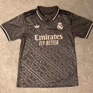 Real Madrid x Adidas svart fotbollströja M - Snygg svart Real Madrid fotbollströja från Adidas i slim fit. Tröjan har korta ärmar, krage med knapp och ett diskret mönster över hela tyget. Tryck med Emirates Fly Better på bröstet och klubbmärke samt Adidas-logga. Materialet är lätt och andas, perfekt för träning eller match.