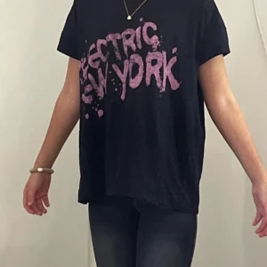 Svart t-shirt med rosa tryck - Cool svart t-shirt med rosa texten 'ELECTRIC NEW YORK' framtill i en splashig stil. Oversized passform och korta ärmar, perfekt till jeans. Troligen i mjuk bomull för skön känsla.