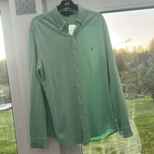 Grön skjorta från Ralph Lauren L - Snygg ljusgrön skjorta från Ralph Lauren i storlek L. Klassisk button-down krage, långa ärmar och knäppning framtill. Broderad röd logga på bröstet.  Bra skick endast använd 3,4 gånger. Säljer pga jag växt ur den. 