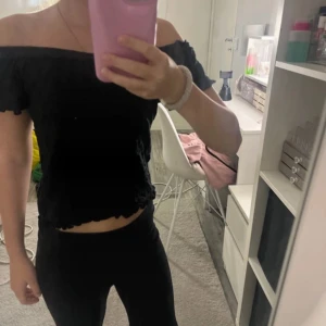 Svart offshoulder croppad topp - Säljer en svart offshoulder topp med croppad passform. Toppen har vågiga kanter vid ärmslut och nederkant, och är helt ärmlös över axlarna. Perfekt för dig som gillar en enkel men snygg stil.