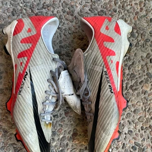Nike Mercurial Air Zoom fotbollsskor - Nike Mercurial Air Zoom fotbollsskor i vitt med svarta och röda detaljer. Pris kan diskuteras . Lite slitna men inte inuti Skorna har snörning, låg profil och mönstrad sula med dobbar för bra grepp på planen. Ovandelen är i syntetmaterial och insidan är röd. Perfekta för dig som vill sticka ut på fotbollsplanen.