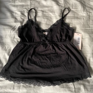 Svart spetslinnetopp från Nelly XS - Supersöt svart linne-topp från Nelly med tunna axelband och v-ringning. Toppen har fina spetsdetaljer längs urringning och nederkant samt en liten rosett framtill. Har aldrig använt linne-toppen då den är lite stor på mig.