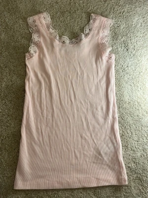 Ljusrosa spetskantad linnetopp Kappahl - Supersöt ljusrosa linnetopp från Kappahl i ribbad bomull med spetsdetaljer och blommönster runt halsringningen och axlarna. Toppen är ärmlös och har en feminin, mjuk look som passar perfekt till jeans eller kjol.