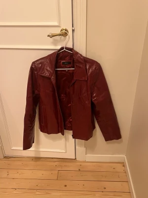 Vinröd skinnjacka med dragkedja - Snygg vinröd skinnjacka från H&M med klassisk krage och raka långa ärmar. Jackan har en stilren look med dragkedja framtill och två fickor på sidorna. Perfekt för dig som vill ha en cool och tidlös jacka i garderoben.