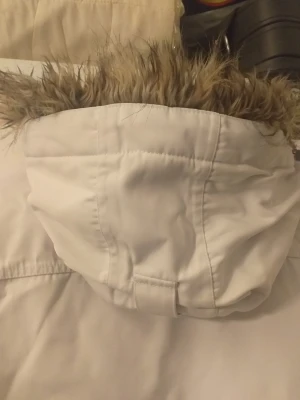 Vit vinterjacka High Mountain strl 40 - Säljer en vit vinterjacka från High Mountain med pälskantad huva och snygga svarta detaljer. Jackan har dragkedja framtill och broderad logga på bröstet. Perfekt för kalla dagar och har en sportig look med coola patches.