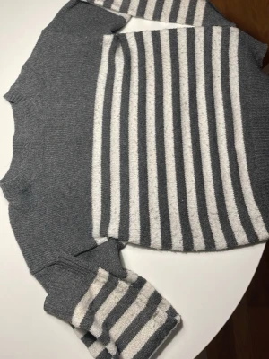Grå och vit randig stickad tröja - Säljer en stickad tröja i grått med vita ränder på både mage och ärmar. Tröjan har rund hals och långa ärmar med breda ribbade muddar. Perfekt för dig som gillar enkel och stilren design med lite edge.