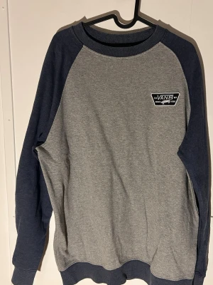 Grå-blå sweatshirt från Vans - Säljer en stilren sweatshirt från Vans i storlek Large. Tröjan är grå med mörkblå ärmar och muddar, samt har en liten Vans-logga på bröstet. Tillverkad i mjuk bomullsmix, perfekt för chill dagar eller när du vill ha en enkel men snygg look.