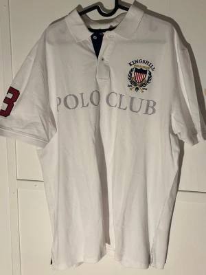 Vit polotopp från Kingshill Polo Club XL - Säljer en vit polotopp från Kingshill Polo Club i storlek XL. Toppen har klassisk krage, korta ärmar och tryckt text 'POLO CLUB' på bröstet samt ett broderat emblem. På ena ärmen finns en stor röd siffra 3. Materialet är bomull och toppen har knappar vid halsen.