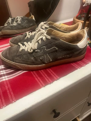 Premiata skor - Säljer ett par grå sneakers från Premiata med vit häl och vita snören. Skorna har en rund tå och är tillverkade i mocka med detaljer i skinn. Snyggt broderad logga på sidan och printad text på hälen. Perfekt för dig som gillar stilrena och klassiska sneakers.