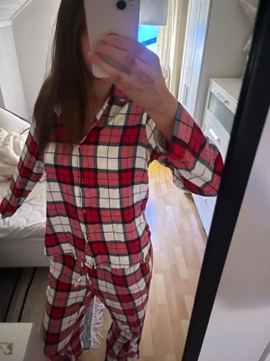Rutig röd och vit pjamas knappar - Säljer en mysig pjamas med klassiskt rutigt mönster i rött, vitt och inslag av mörkblått. Pyjamasen har krage och knäppning framtill med röda knappar. Tröjan har storlek: xs och byxan: m.
