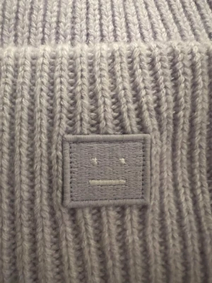 Ljuslila stickad mössa Acne Studios - Säljer en ljuslila stickad mössa från Acne Studios med det klassiska ansikts-emblemet framtill. Mössan är ribbstickad och har en bred uppvikt kant. Perfekt för kyliga dagar 