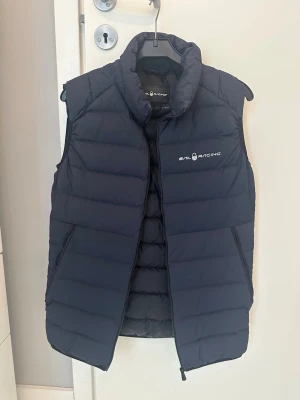 Mörkblå dunväst från Sail Racing - Säljer en mörkblå dunväst från Sail Racing med hög krage och vit logotyp på bröstet och ryggen. Västen har quiltad design och dragkedja framtill samt två sidofickor med dragkedja. Perfekt för lager-på-lager under kyliga dagar.