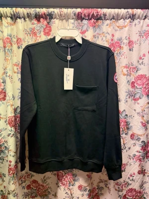 Svart sweatshirt från Canada Goose L - Svart sweatshirt från Canada Goose i storlek L. Tröjan har en rund hals, en bröstficka och ett broderat märke på ärmen. Materialet är en mix av polyester och bomull, vilket gör den mjuk och skön. Perfekt för dig som gillar stilrena och klassiska plagg.