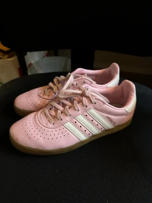 Adidas 350 rosa sneakers med snörning - Säljer ett par Adidas 350 sneakers i ljusrosa skinn med klassiska vita ränder på sidorna och beige gummisula. Skorna har perforerat mönster på tån och snörning framtill. Perfekt för dig som gillar sportig och clean stil.