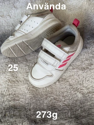 Vita adidas sneakers med rosa detaljer - Säljer ett par vita adidas sneakers med rosa ränder och praktiska kardborreband. Skorna har rund tå, platt sula och en rosa hälflik. Perfekta för dig som gillar sportig stil och enkel på- och avtagning.🌸 Använt skick. Hela. Ej tvättade. Stl 25
