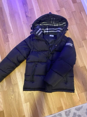 Svart pufferjacka från Burberry - Svart pufferjacka från Burberry med avtagbar huva och klassiskt rutigt foder i grått och vitt. Jackan har dragkedja, tryckknappar och två fickor framtill. På vänster ärm finns en tydlig Burberry London England-logga.