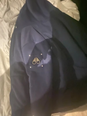 Mörkblå pufferjacka från Moose Knuckles - Säljer en mörkblå pufferjacka från Moose Knuckles med ikonisk logga på ärmen och quiltad insida med mönster. Jackan har dragkedja och är fodrad med polyester för extra värme. Perfekt för dig som vill ha en snygg och varm jacka med streetkänsla.