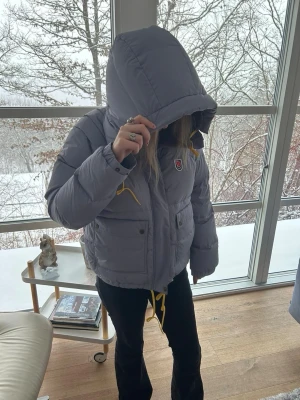 Ljusgrå pufferjacka från Fjällräven - Säljer en ljusgrå pufferjacka från Fjällräven åt tjejen  med huva och gula snören. Jackan har två stora fickor med knappar framtill, dragkedja och ett klassiskt Fjällräven-märke på bröstet. Perfekt för kalla dagar och har en schysst, lite boxig passform.