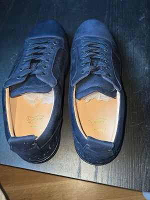 Mörkblå sneakers från Christian Louboutin - Säljer ett par mörkblå sneakers från Christian Louboutin med lyxig mocka och signaturröd sula. Skorna har snörning, rund tå och diskret broderad logga på sidan. Tillverkade i Italien med snygga detaljer och platt sula. Perfekt för dig som gillar exklusiva sneakers.