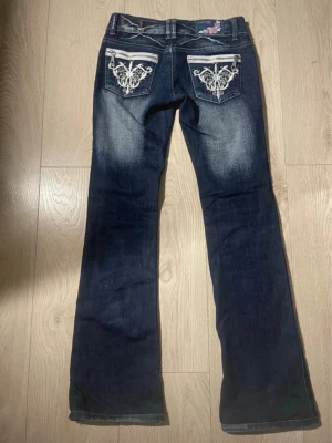 Blå bootcut jeans från Crazy Age - Säljer ett par mörkblå bootcut jeans från Crazy Age med snygga broderade detaljer på bakfickorna och rosa broderad logga vid linningen. Jeansen har låg midja, klassisk femficksdesign och slitningar framtill för en cool look.