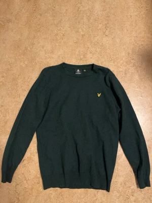 Mörkgrön stickad tröja Lyle & Scott - Mörkgrön stickad tröja från Lyle & Scott med klassisk rund hals och gul broderad logga på bröstet. Tröjan har långärmad passform och ribbade muddar vid ärmslut och nederkant. Perfekt för dig som gillar stilrena och enkla plagg.