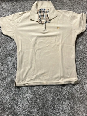 Beige kortärmad Burberry Pike XL - Säljer en beige kortärmad Piké från Burberry i storlek XL. Klassisk krage med rutigt Burberry-mönster på insidan och ärmslut, samt liten gul broderad logga på bröstet. Toppen har normal passform och är tillverkad i mjuk bomull.Nypris 4000