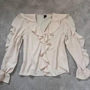 Beige volangblus från Vero Moda XS - Supersöt beige blus från Vero Moda i storlek XS. Blusen har långa ärmar med volangdetaljer och v-ringning framtill med volanger längs halsringningen. Tillverkad i ett lätt och flowy material.