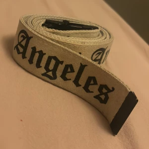 Beige canvasbälte med Los Angeles-tryck - Säljer ett vitt canvasbälte med svart text i gotisk stil där det står 'Los Angeles'. Bältet har ett svart metallspänne som ger en cool kontrast mot det ljusa tyget. Perfekt accessoar för att lyfta din outfit med streetkänsla.