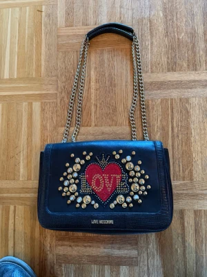 Svart axelväska från Love Moschino - Snygg svart axelväska från Love Moschino med guldfärgade kedjor och detaljer. Framsidan har ett stort rött hjärta med texten 'LOVE' och glittriga nitar runt om. Insidan är fodrad i rött tyg med logotryck och har en dragkedjeficka. Perfekt för dig som gillar statement-accessoarer.