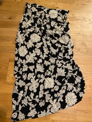 Svart långkjol med blommigt mönster - Säljer en svart asymmetrisk kjol med vita blommor och paisley-inspirerat mönster. Kjolen har en avslappnad passform och är perfekt för dig som gillar bohemisk stil. Materialet känns mjukt och luftigt, vilket gör den skön att bära under varmare dagar.