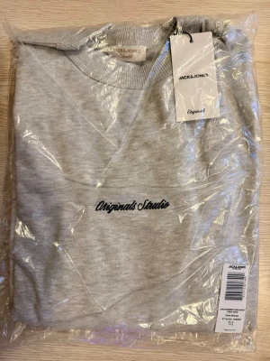 Grå sweatshirt från Jack & Jones - Säljer en stilren ljusgrå sweatshirt från Jack & Jones Originals Studio. Tröjan har rund hals, broderad logga på bröstet och är tillverkad i mjuk bomullsmix. Perfekt för dig som gillar enkel och clean streetwear.