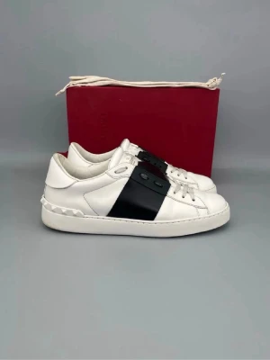 Valentino Garavani vita sneakers - Säljer ett par vita Valentino Garavani sneakers med bred svart rem över mitten och coola nitar på hälen. Skorna har snörning och är tillverkade i skinn för en lyxig känsla. Perfekta för dig som vill ha en clean men ändå edgy look.