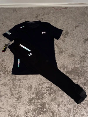 Under armour reflektiv set tracksuit - Bilderna visar ett svart träningsset från Under Armour med t-shirt och byxor, båda med färgglada loggor och detaljer. 