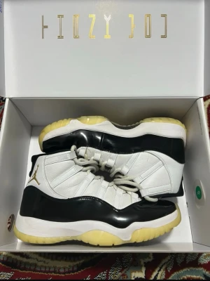 Jordan 11 Retro DMP Defining Moments 11 svart/vit - Nike Air Jordan 11 sneakers i svart och vit med gulddetaljer. Skorna har högt skaft, snörning och en platt sula. Ovandelen är i skinn och blankt syntetmaterial, med rund tå och guldfärgad Sulan är gulaktig och ger en retro vibe.